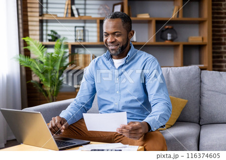 Smiling man enjoying remote work from home office while looking at financial documents. Entrepreneur managing business finances using laptop at home in cozy living room setting 116374605