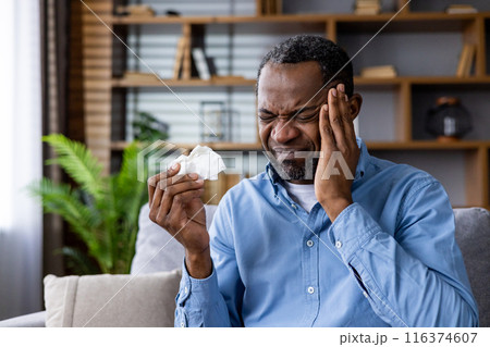 Man in blue shirt holding tissue with pained expression indicating headache. Home setting with bookshelves and plants in background. Concept of sickness, stress, and discomfort. 116374607