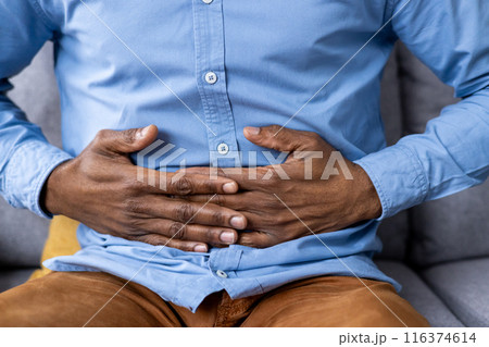 Close-up of man's hands clutching stomach indicating abdominal pain and discomfort. Concept of digestive issues, stomach ache, and health problems. Man in blue shirt and brown pants sitting on couch. Close-up of man's hands clutching stomach indicating abdominal pain and discomfort. Concept of digestive issues, stomach ache, and health problems. Man in blue shirt and brown pants sitting on couch. 116374614