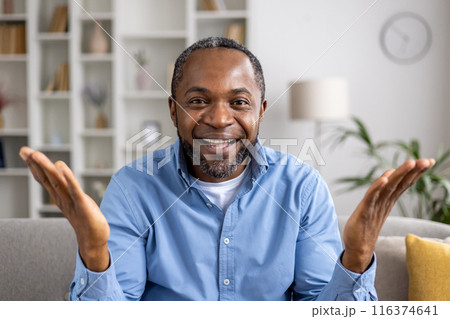 Happy man smiling and looking into camera during video call. Comfortable home setting with bookshelves, lamp, and indoor plants. Concept of communication, happiness, and technology. 116374641