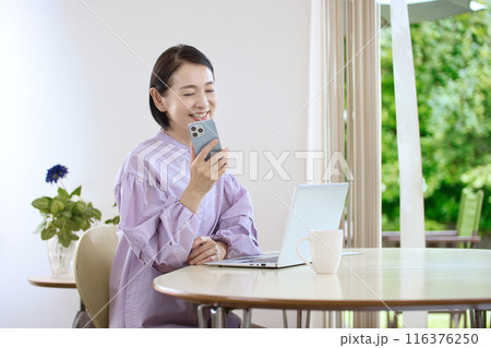 リビングでスマホを操作しながらパソコンを見るミドル女性 116376250