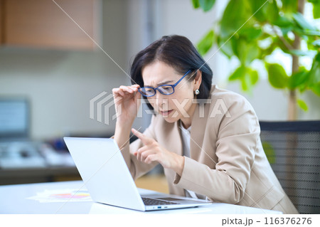 老眼鏡を掛けてオフィスのパソコンを見るミドル女性        116376276