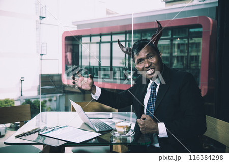 Man in suit with horns sitting at desk with laptop and documents, gesturing expressively, with cup of coffee and cafe background Man in suit with horns sitting at desk with laptop and documents, gesturing expressively, with cup of coffee and cafe background 116384298