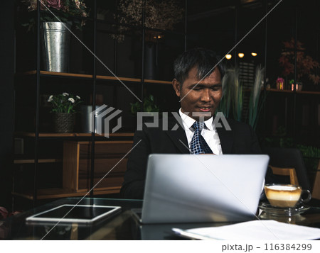 Smiling Businessman in suit working on laptop at dark, modern office desk with tablet and cup of coffee 116384299