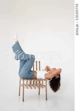 Happy beautiful girl posing with a chair in a new hot look. Wearing blue jeans and white waistcoat Happy beautiful girl posing with a chair in a new hot look. Wearing blue jeans and white waistcoat 116385759