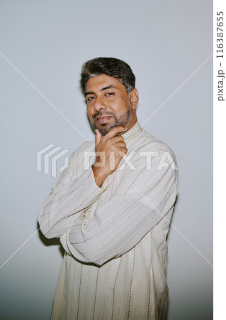 Portrait of young Indian man dressed in patterned creamy shirt holding his chin and smirking with squint while sitting for photo Portrait of young Indian man dressed in patterned creamy shirt holding his chin and smirking with squint while sitting for photo 116387655