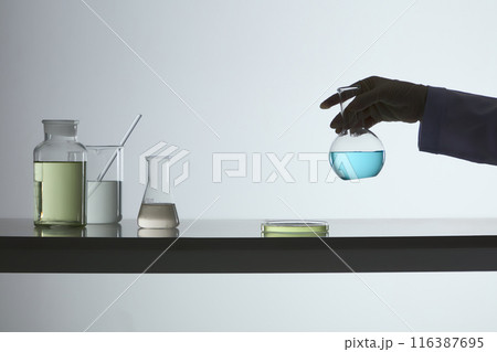A scientist hand holding a boiling flask of blue fluid, the table featuring some glassware in the experiment, against the white background. The photo was taken from front angle, vacant space A scientist hand holding a boiling flask of blue fluid, the table featuring some glassware in the experiment, against the white background. The photo was taken from front angle, vacant space 116387695