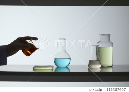 A beaker of yellow fluid held by a scientist hand, prepare to pour into a petri dish, which displayed on the table next to a boiling flask, erlenmeyer flask and glass bottle. Copy space for text 116387697