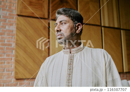 Low angle shot of adult Indian man dressed in creamy flax shirt standing still and looking away Low angle shot of adult Indian man dressed in creamy flax shirt standing still and looking away 116387707