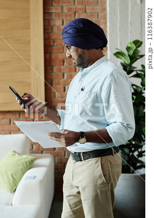 Adult Indian worker holding recent research while checking his phone at same time 116387902