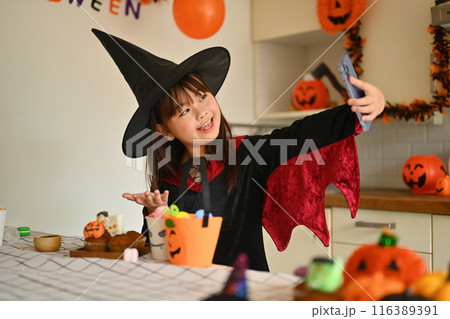 Smiling little girl in witch costumes taking selfies with decorated Halloween cupcakes in the kitchen 116389391