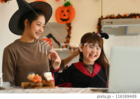 Mother and her daughter dressed in witch costumes having video call on laptop in kitchen. 116389489
