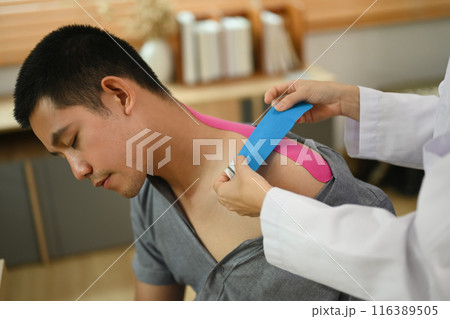 Physical therapist placing pink kinesio tape on man patient shoulder and neck at the clinic office. Alternative medicine concept. 116389505