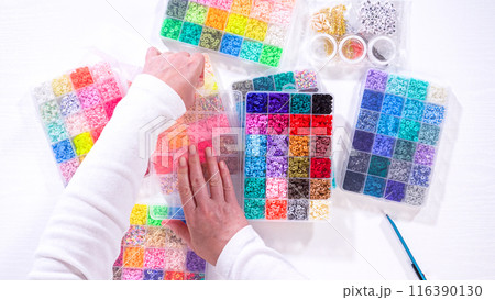 Flat lay. Woman hands gracefully poised over a collection of beads, sorted by color in transparent organizers. The array of beads spans a vibrant spectrum, from deep purples to bright oranges 116390130