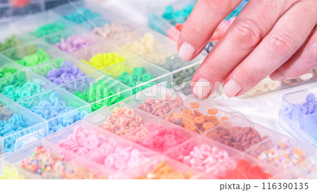 Woman hands gracefully poised over a collection of beads, sorted by color in transparent organizers. The array of beads spans a vibrant spectrum, from deep purples to bright oranges, meticulously Woman hands gracefully poised over a collection of beads, sorted by color in transparent organizers. The array of beads spans a vibrant spectrum, from deep purples to bright oranges, meticulously 116390135