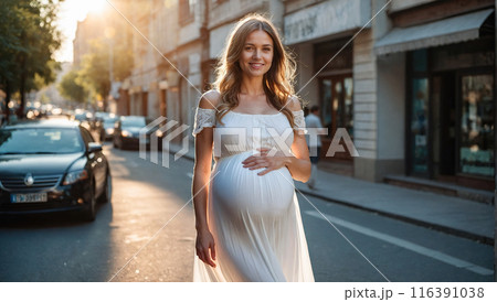 A smiling pregnant woman in a white dress walking down a sunlit street, radiating joy and anticipation. 116391038