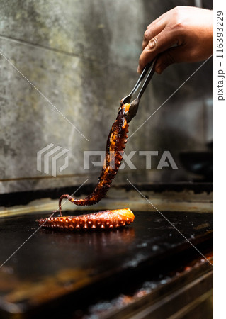 Hand of a cook catching an octopus tentacle with the tongs on a restaurant grill. Vertical Hand of a cook catching an octopus tentacle with the tongs on a restaurant grill. Vertical 116393229