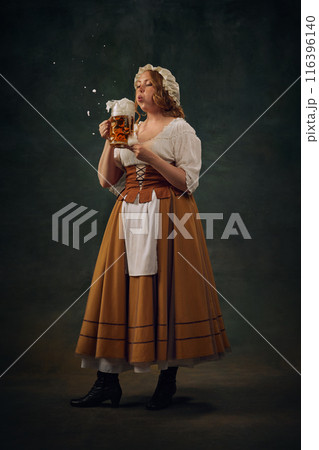 Woman in historical attire, wearing bonnet, white blouse, and brown skirt with apron, holding frothy beer mug and blowing foam against vintage background. Woman in historical attire, wearing bonnet, white blouse, and brown skirt with apron, holding frothy beer mug and blowing foam against vintage background. 116396140