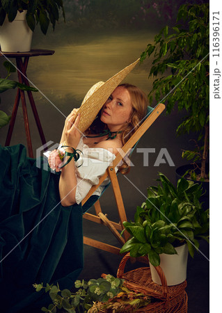 Woman in historical attire, wearing large straw hat, white blouse, and green velvet skirt, sitting on chair, partially hiding her face with hat, surrounded by potted plants. 116396171