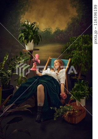 Super hot days. Woman in 18s century attire sitting tired in chair in green garden with small pink electric fan blowing on her, against vintage green background 116396199