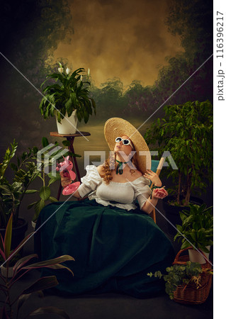 Portrait of elegant woman in period attire sitting on chair in green garden with small pink electric fan blowing on face, against vintage background. Hot summer day picnic gathering 116396217