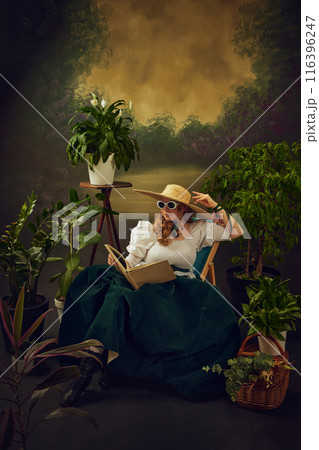 Portrait of elegant young woman in retro style glasses, straw hat, and velvet skirt, sitting on chair around lush green plants, and reading book against vintage background Portrait of elegant young woman in retro style glasses, straw hat, and velvet skirt, sitting on chair around lush green plants, and reading book against vintage background 116396247