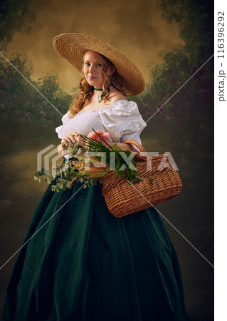 Portrait of woman in historical clothes, in large straw hat, white blouse, and green velvet skirt, holds wicker basket filled with greenery and flowers, set against vintage background Portrait of woman in historical clothes, in large straw hat, white blouse, and green velvet skirt, holds wicker basket filled with greenery and flowers, set against vintage background 116396292