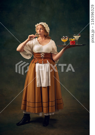 Portrait of woman in historical attire, drinking cocktail and holding a tray with various alcoholic drinks, set against vintage green background 116396329