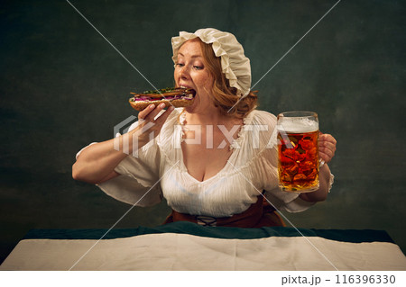 Young woman in medieval clothes taking giant bite of sandwich with fish and onion, holding beer mg, sitting at against vintage green background 116396330