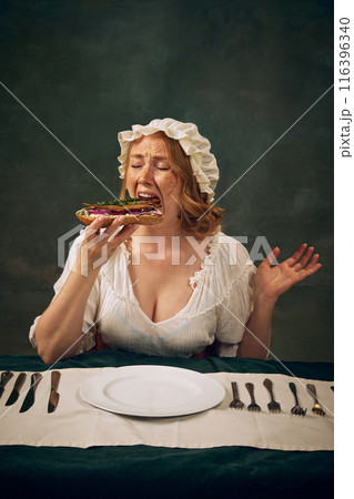 Portrait of woman in period costume taking large, enthusiastic bite of sandwich with fish and onions, sitting at table with plate, knives and forks, set against vintage background 116396340