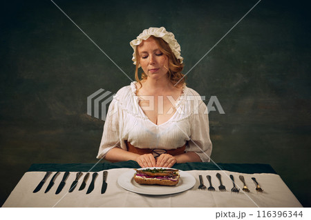 Portrait of beautiful young woman in bonnet and white blouse, sitting at table with multiple knives and forks, looking on late with delicious bruschetta with fish and onion 116396344