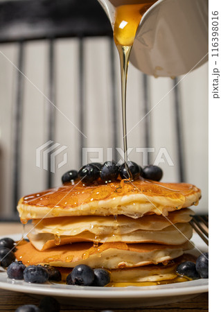 Stack of homemade pancakes prepared with blueberries and a jar with honey falling on the pancakes an old plate on a wooden table. Concept healthy food at breakfast. 116398016