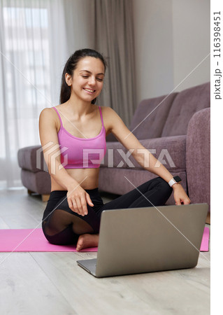 Happy Caucasian woman looking at laptop while sitting on pink fitness mat, ready for online training and workout at home. Online training. 116398451