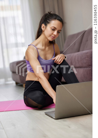 Happy sporty Caucasian woman looking at laptop while sitting on pink fitness mat, ready for online training and workout at home. 116398455