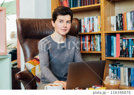 Confident middle aged female expert sits with laptop in leather armchair in cozy office with bookshelf background. 40s middle-age professional portrait of teacher, coach, mentor, therapist, counselor. Confident middle aged female expert sits with laptop in leather armchair in cozy office with bookshelf background. 40s middle-age professional portrait of teacher, coach, mentor, therapist, counselor. 116398878