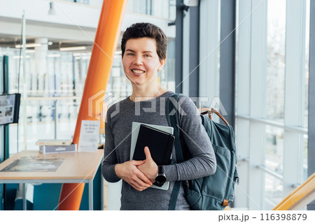 Smiling neutral gender middle-aged female person in casual clothes with backpack and books stands on staircase in modern building of university or education Centre. Adult students, Academic education. Smiling neutral gender middle-aged female person in casual clothes with backpack and books stands on staircase in modern building of university or education Centre. Adult students, Academic education. 116398879