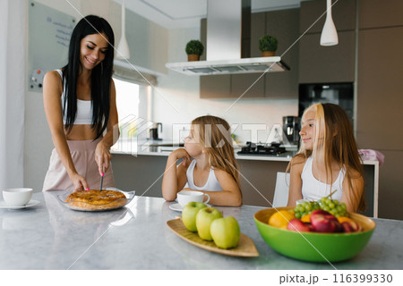 Mom cuts a piece of apple pie for her children daughters who are sitting at the dining table for breakfast in their pajamas. 116399330