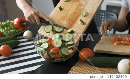 Mother with her daughter are preparing a fresh salad together, slicing cucumbers and bell peppers, adding ingredients to a bowl 116402569