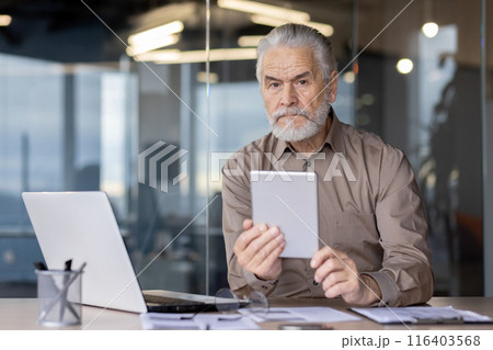 Senior businessman with beard using tablet seated at modern office desk with laptop, documents, and office supplies. Focused expression, professional attire, contemporary background 116403568