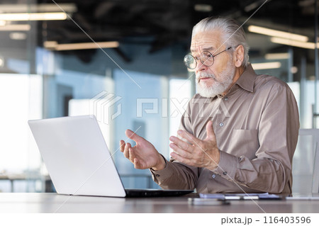Elderly man using laptop in bright, modern office participating in video conference call. Expressive hand gestures suggesting conversation or presentation. Professional setting with open workspace 116403596