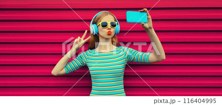 Stylish young woman taking selfie with smartphone listening to music with headphones using phone Stylish young woman taking selfie with smartphone listening to music with headphones using phone 116404995