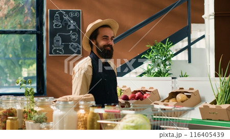 Happy man spraying fresh produce to preserve aroma and smell, zero waste supermarket vendor. Owner with hat and apron taking care of fruits and vegetables placed for sale, neighborhood shop. 116405439