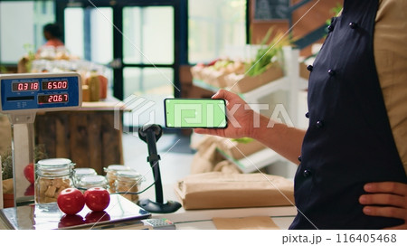 Merchant with phone greenscreen at store counter, presenting isolated chromakey display on mobile gadget. Farmer and vendor using modern copyspace template with mockup technology. 116405468