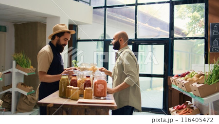 Shop owner shows pasta types to client, opening reusable containers filled with bulk items in natural bio grocery store. Vendor recommending organic additives free food goods. 116405472