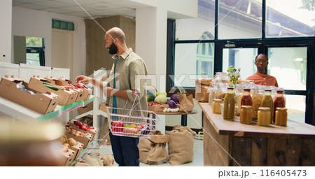 Buyer choosing ripe bio vegetables to buy for healthy sustainable dinner, looking at organic homegrown produce in crates at zero waste eco store. Man examining natural groceries. Tripod shot. 116405473