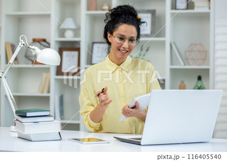 Smiling woman enjoying a study session at home. She is taking notes and using a laptop for online education. The cozy home office setup includes books, a lamp, and a tablet Smiling woman enjoying a study session at home. She is taking notes and using a laptop for online education. The cozy home office setup includes books, a lamp, and a tablet 116405540