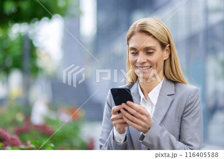 Confident businesswoman in gray suit smiling while using smartphone outdoors. Professional woman engaged in communication and networking through mobile device in city environment. Confident businesswoman in gray suit smiling while using smartphone outdoors. Professional woman engaged in communication and networking through mobile device in city environment. 116405588