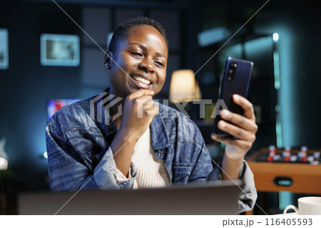 Close-up of smiling black woman grasping and looking at her mobile device. Portrait of african american female influencer using smartphone to browse the internet and surf social media. Close-up of smiling black woman grasping and looking at her mobile device. Portrait of african american female influencer using smartphone to browse the internet and surf social media. 116405593