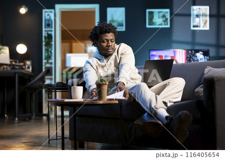 BIPOC man in apartment paying attention in elearning seminar teleconference with teacher, writing on notepad. Homeschooling student listening to lesson and checking presentation files 116405654