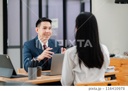 Two professionals engaged in a business meeting, discussing work at a modern office with laptops and documents on the table. Two professionals engaged in a business meeting, discussing work at a modern office with laptops and documents on the table. 116410978